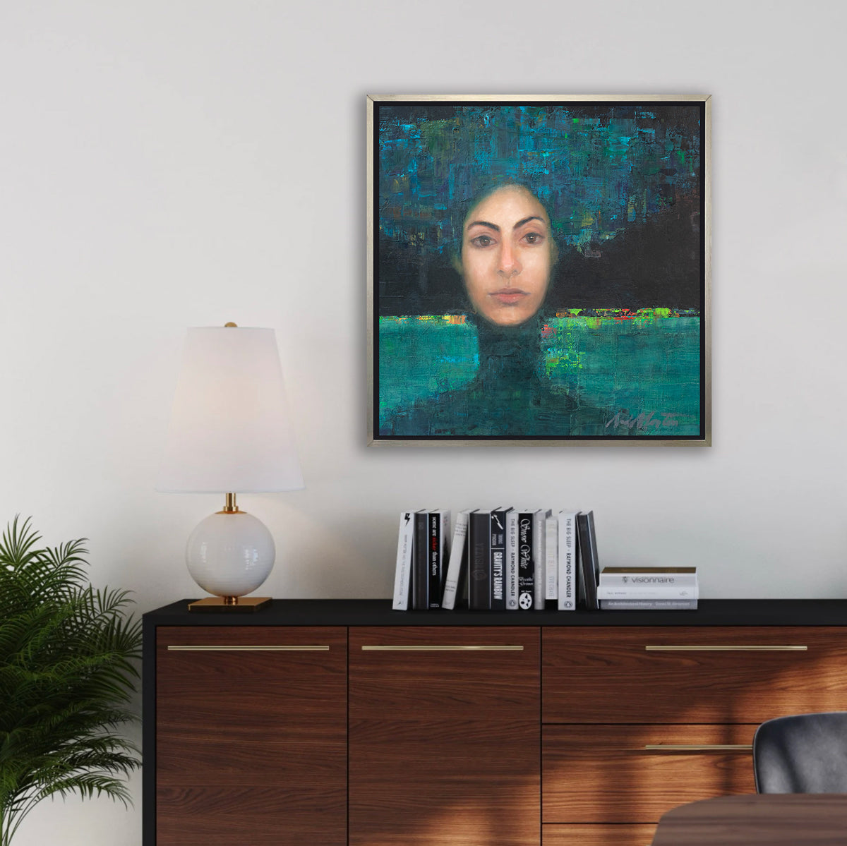 A blue, black, teal and beige abstract figural print in a silver floater frame hangs on a white wall above a black and dark wood cabinet with a stack of books and a white table lamp.