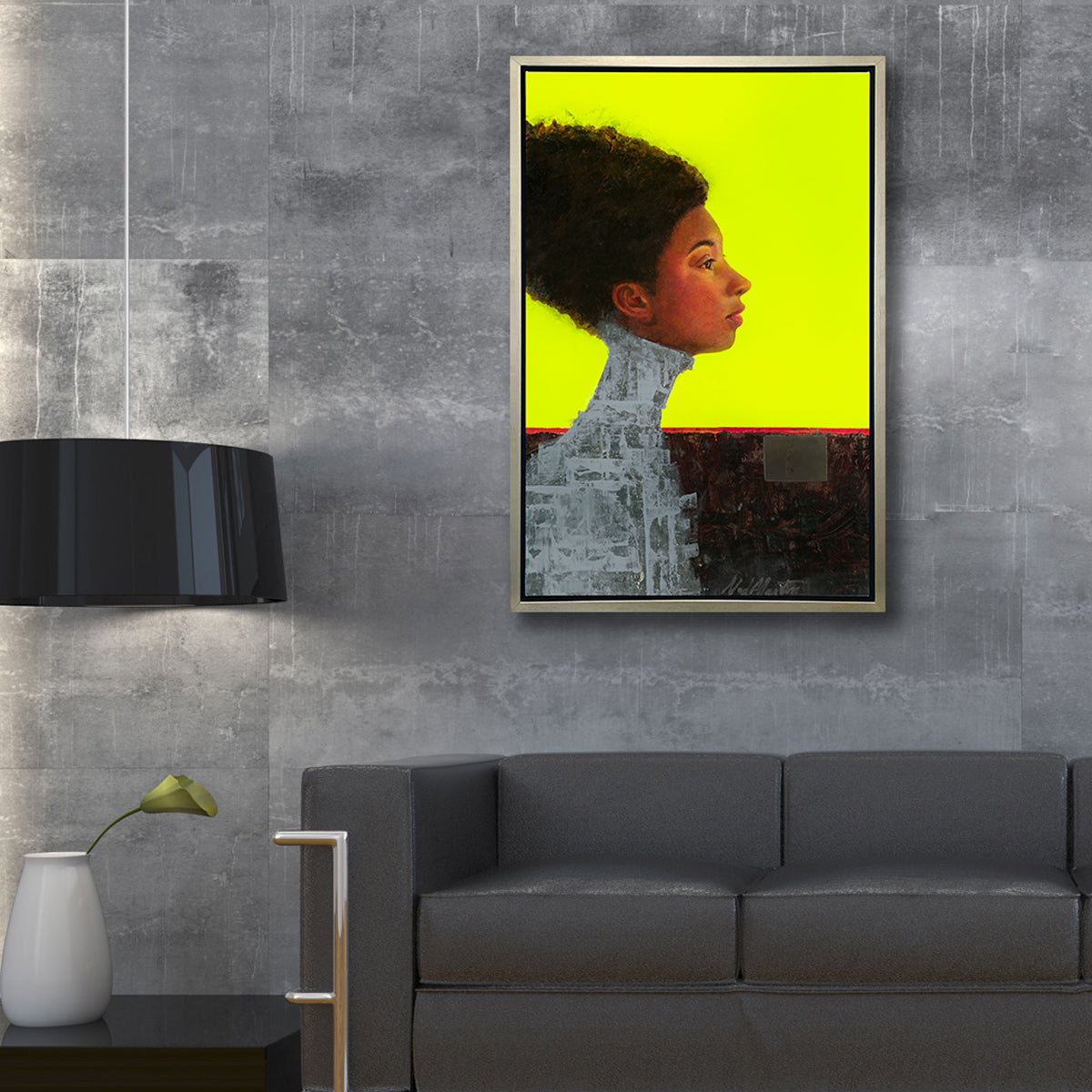 A grey, brown red and neon yellow abstract figural print in a silver floater frame hangs on a concrete wall above a dark grey sofa, a dark wood side table with a white vase with a green flower and black shade lamp pendant.