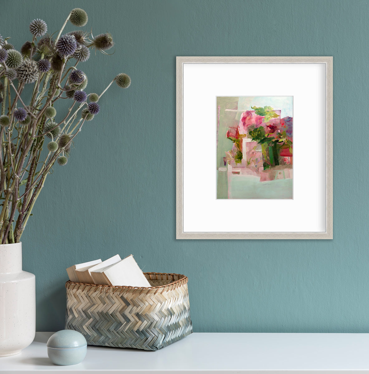 A pink, green and red abstract print in a silver frame with a mat hangs on a teal wall above a white surface with a woven beige and teal basket, a light teal trinket and a white vase with green and purple pom pom stems.