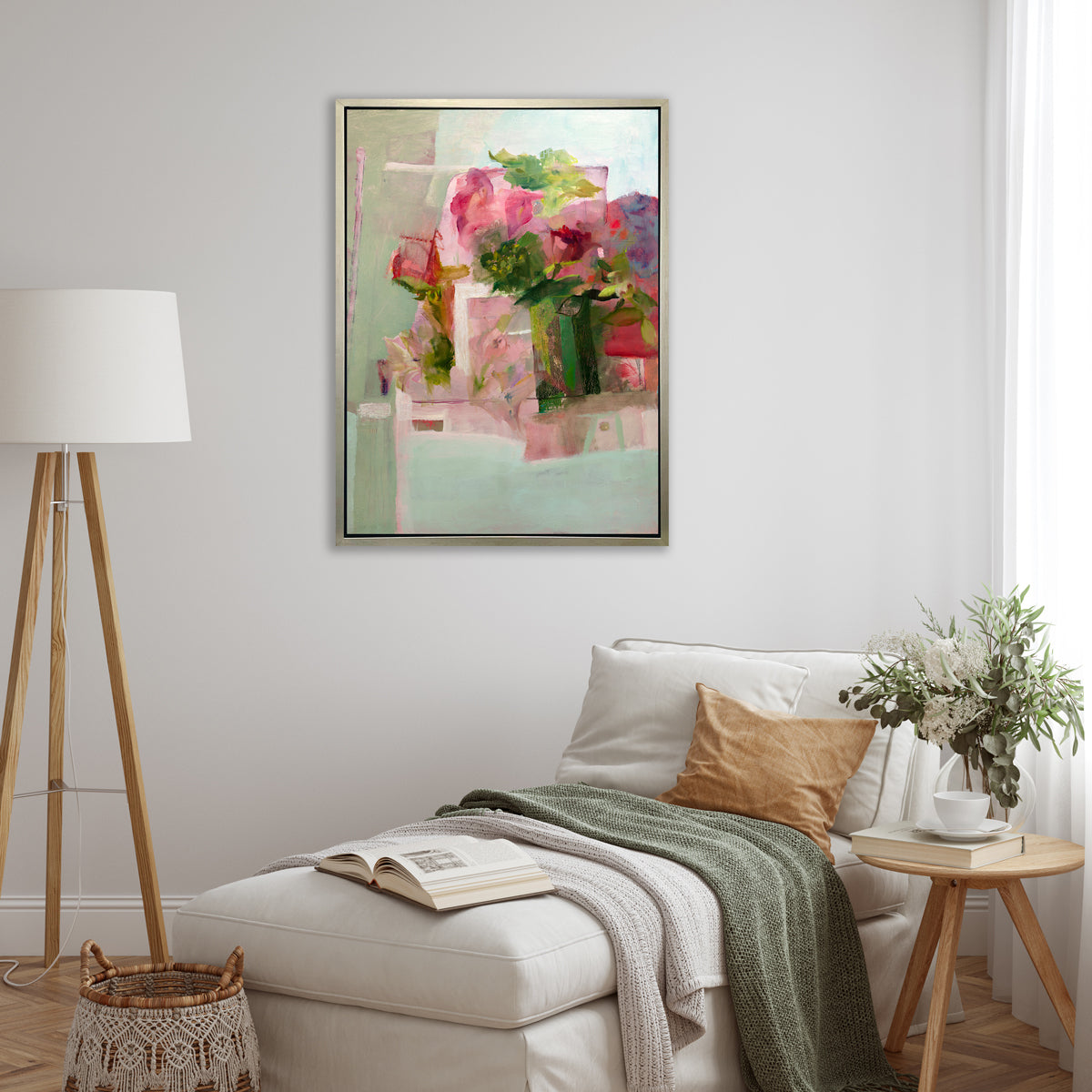 A pink, green and red abstract print in a silver floater frame hangs on a white wall above a white lounge chair with pillows and a green throw blanket, a round wood side table with a floral arrangement and a book and a tripod floor lamp in the corner.