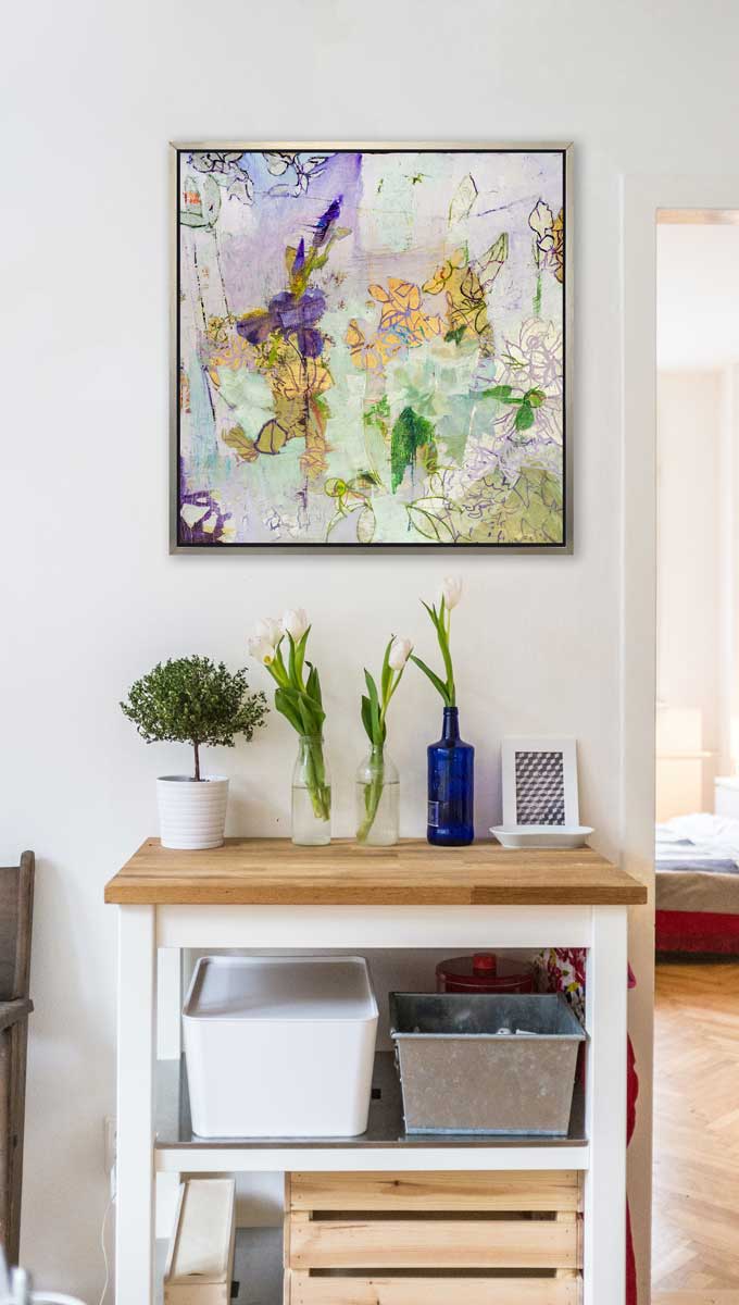 A purple, green and gold abstract print in a silver floater frame hangs on a white wall above a butcher block and white island cart with rectangular storage containers, a small potted shrub and bottle vases with white tulips.