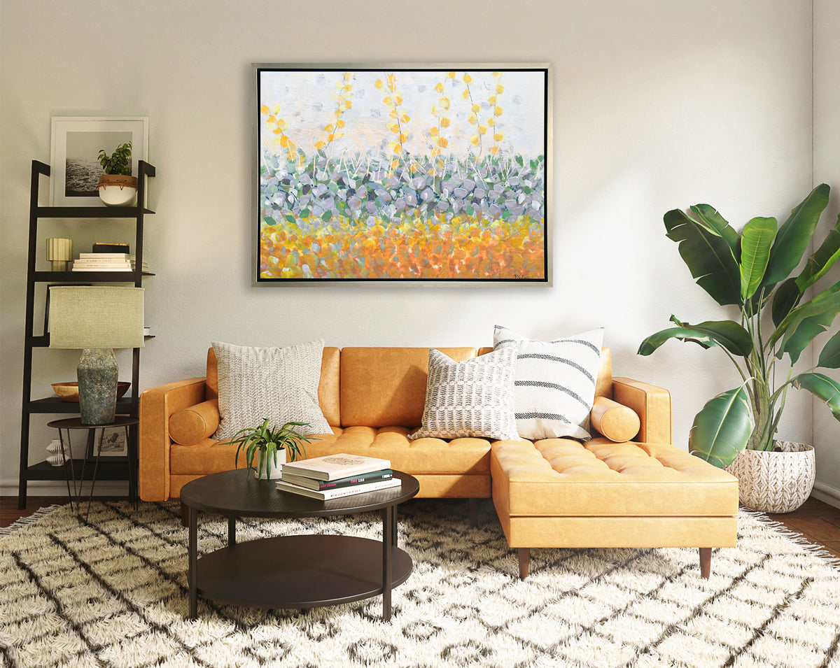 A yellow, purple, green and orange abstract print in a silver floater frame hangs on a white wall above a orange sofa with neutral pillows, a dark wood round coffee table with books and a plant, a tall bookshelf with decorative accessories and a potted floor plant.