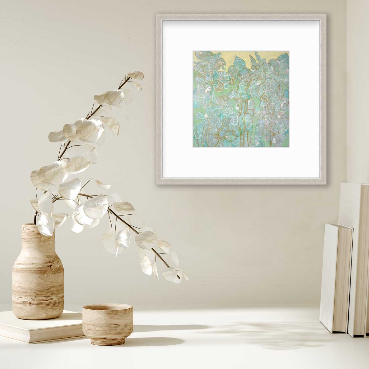 A green, yellow and white abstract print in a silver frame with a mat hangs on a white wall above a couple books and a wood vase with white leaf stems.