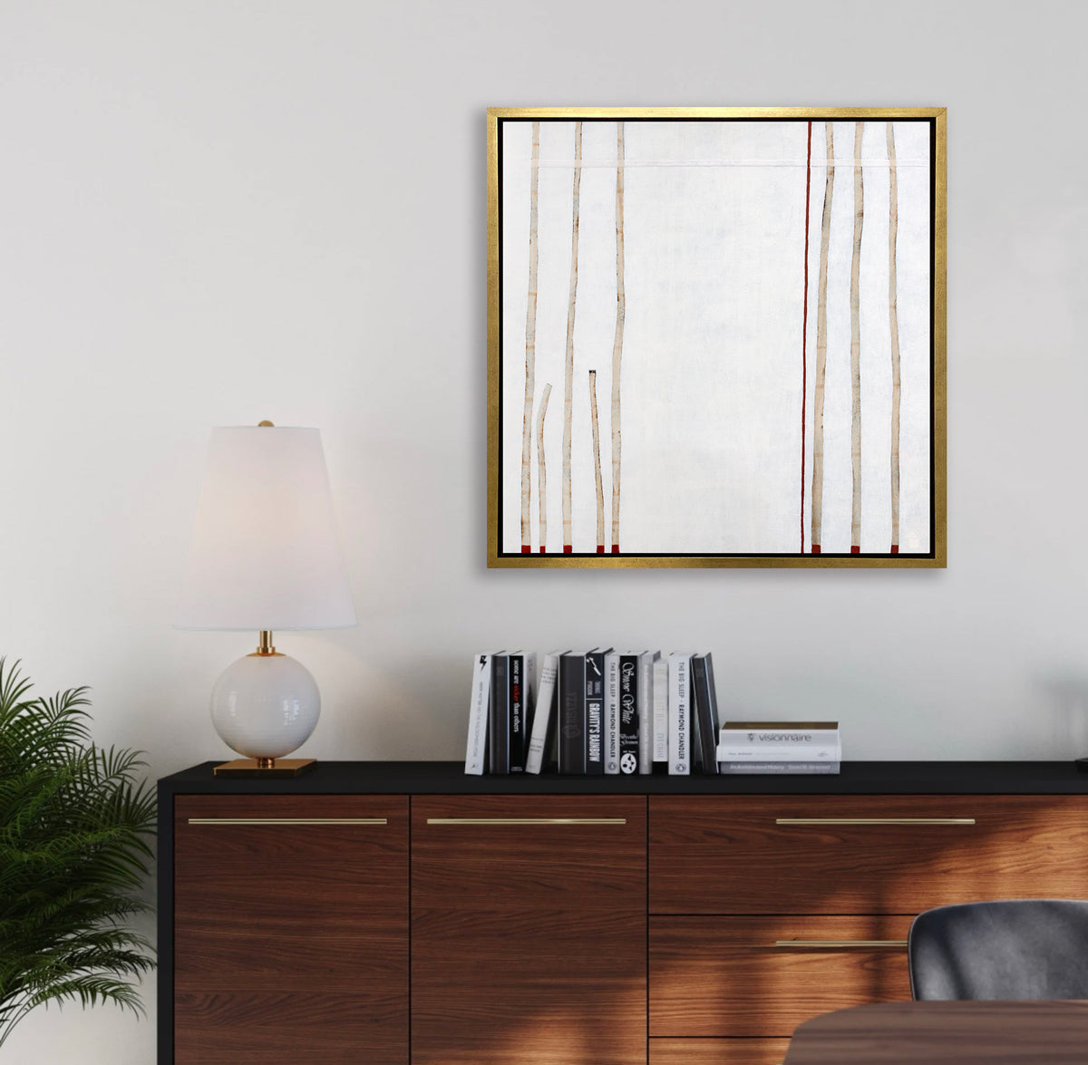 A white, beige and red abstract print in a gold floater frame hangs on a white wall above a dark wood and black console cabinet with a stack of books and a white table lamp.
