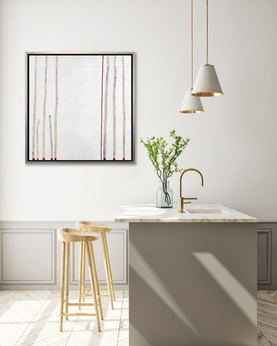 A white, beige and red abstract print in a silver floater frame hangs on a white wall above a white marble countertop kitchen island with a sink and a bottle with green leaf stems and two wood counter stools.