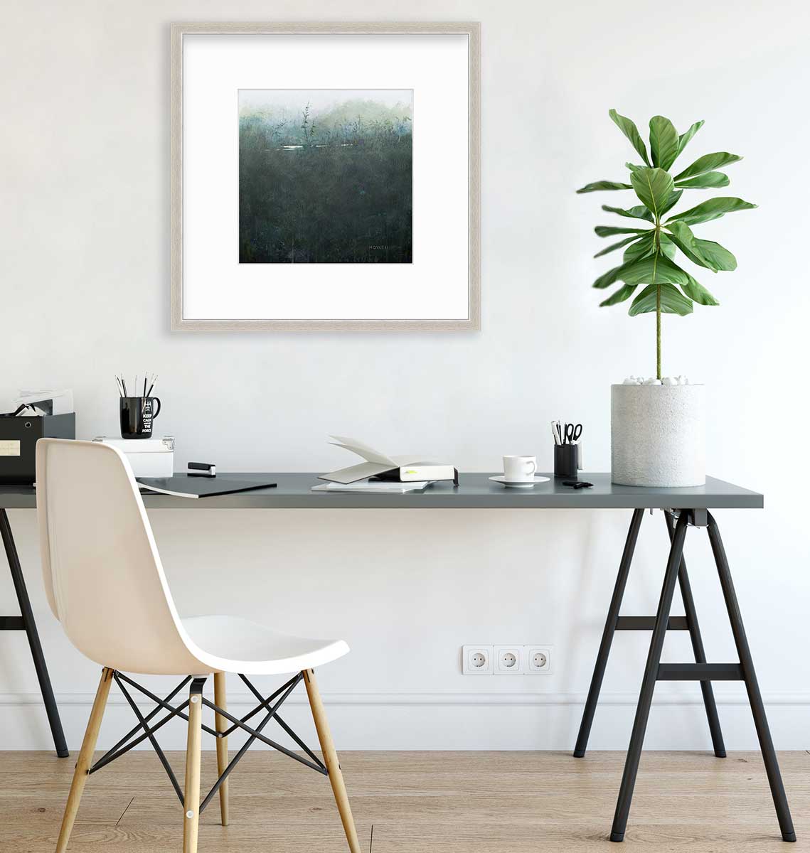 A dark muted teal abstract landscape print in a silver frame with a mat hangs on a white wall above a dark grey desk with open notebooks, a black coffee mug with pencils and a potted plant.