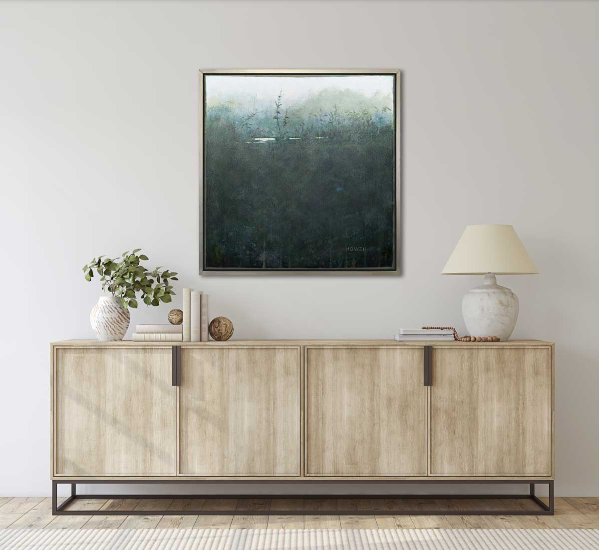 A dark muted teal abstract landscape print in a silver floater frame hangs on a white wall above a wooden sideboard cabinet with a white table lamp, a stack of books and a vase with eucalyptus stems.