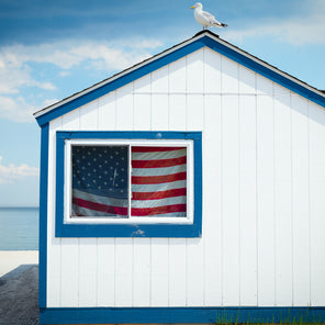 A photograph of white and blue building on the water with an American flag in the window and a seagull resting on the roof in Westport, Connecticut. 