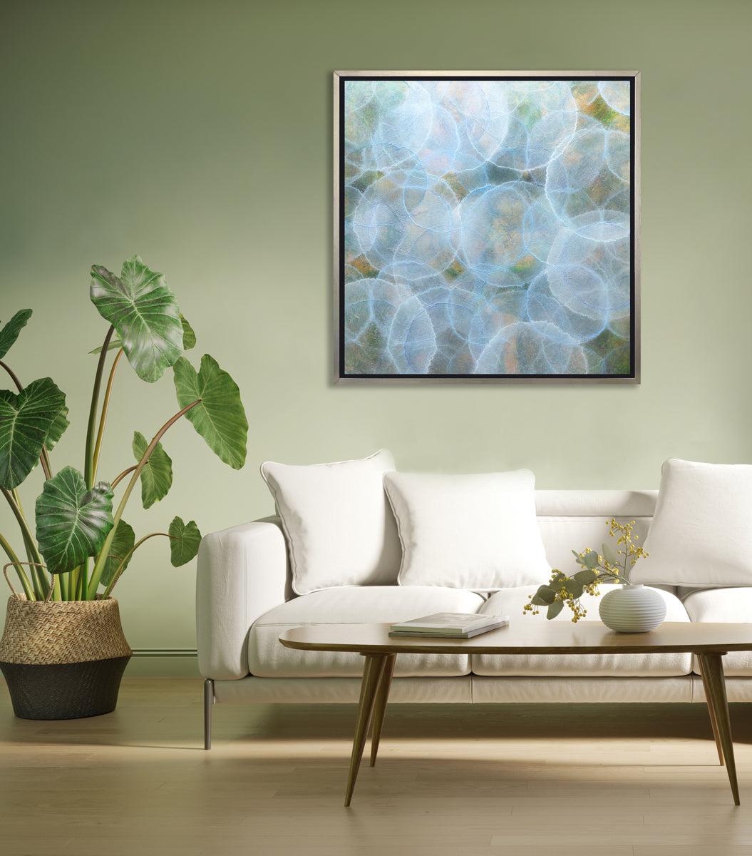 A light blue, green and yellow abstract geometric print in a silver floater frame hangs on a light green wall above a white sofa with pillows, a oval wood coffee table with decorative accessories and a potted floor plant.