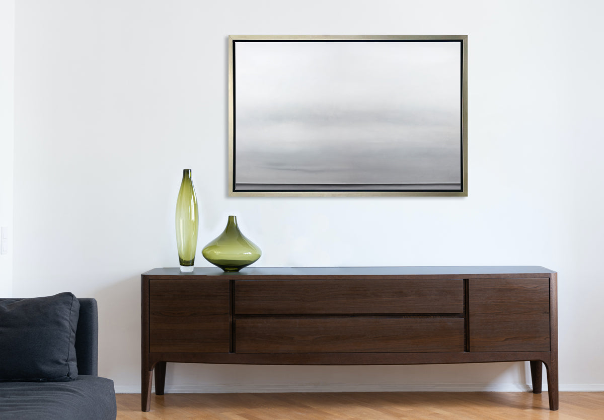 A tonal grey abstract landscape print in a silver floater frame hangs on a white wall above a dark wood sideboard with two green vases in different shapes and sizes.