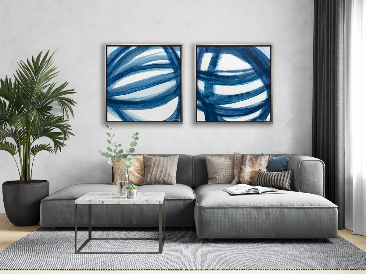 Two blue and white abstract prints in silver floater frames hang side by side on a white wall above a grey oversized sectional sofa with neutral pillows, a square white marble coffee table with decorative accents and a large potted floor plant.