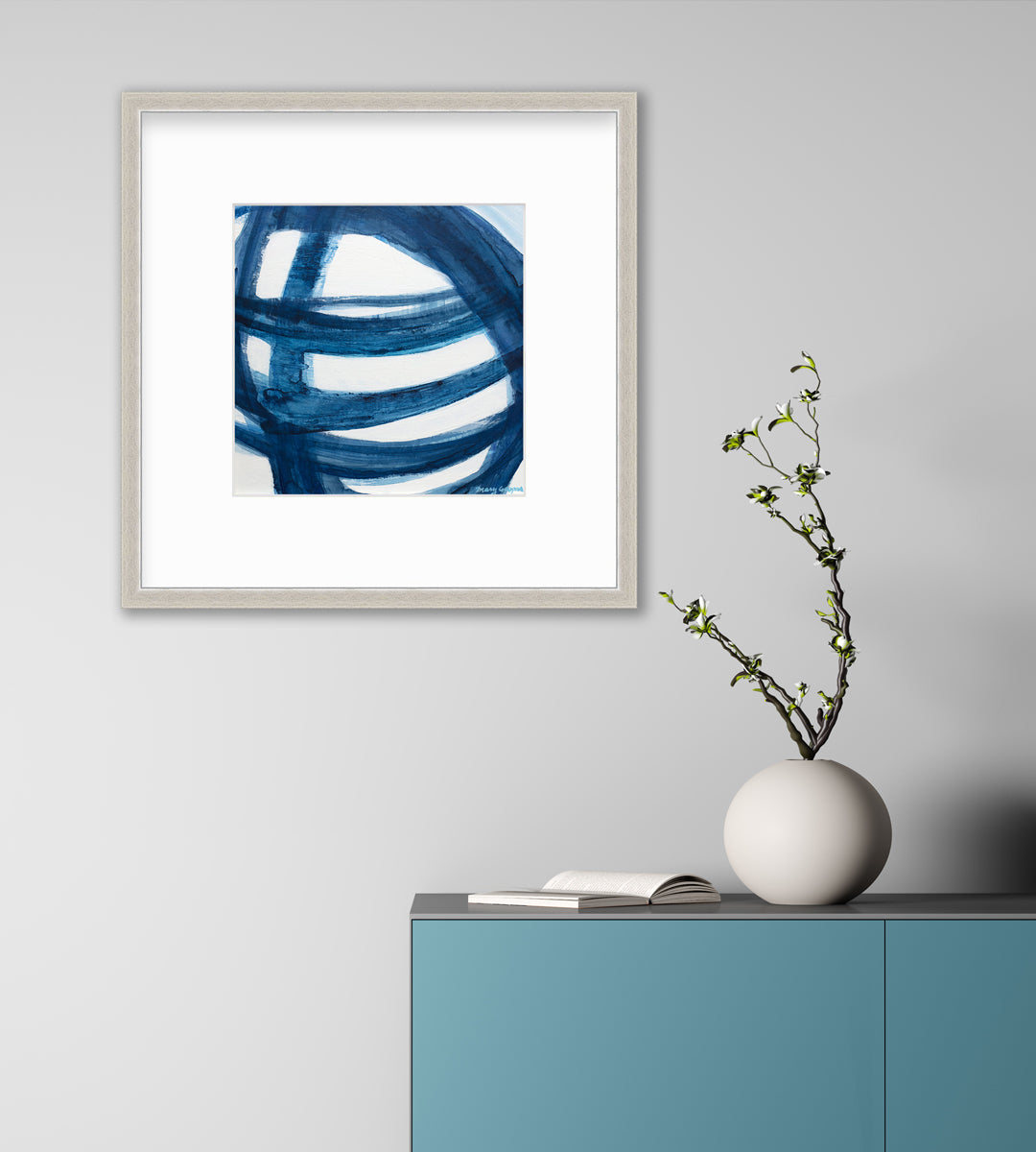 A blue and white abstract print in a silver frame with a mat hangs on a white wall above a teal cabinet with an open book and a white rounded vase with greenery stems.