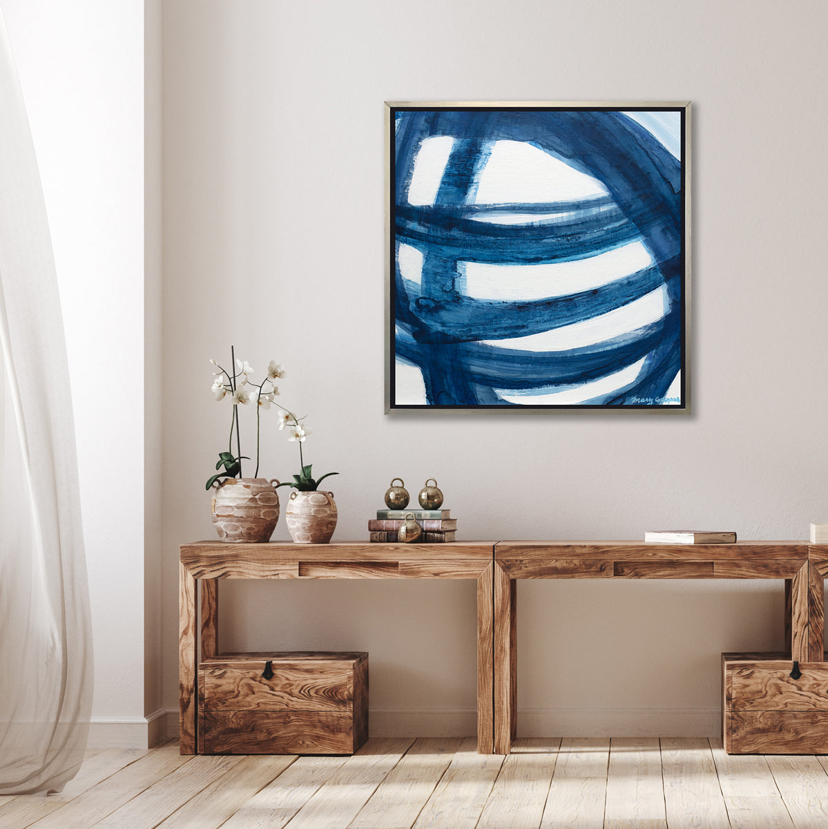 A blue and white abstract print in a silver floater frame hangs on a white wall above a wood desk with a stack of books and two potted white orchids.