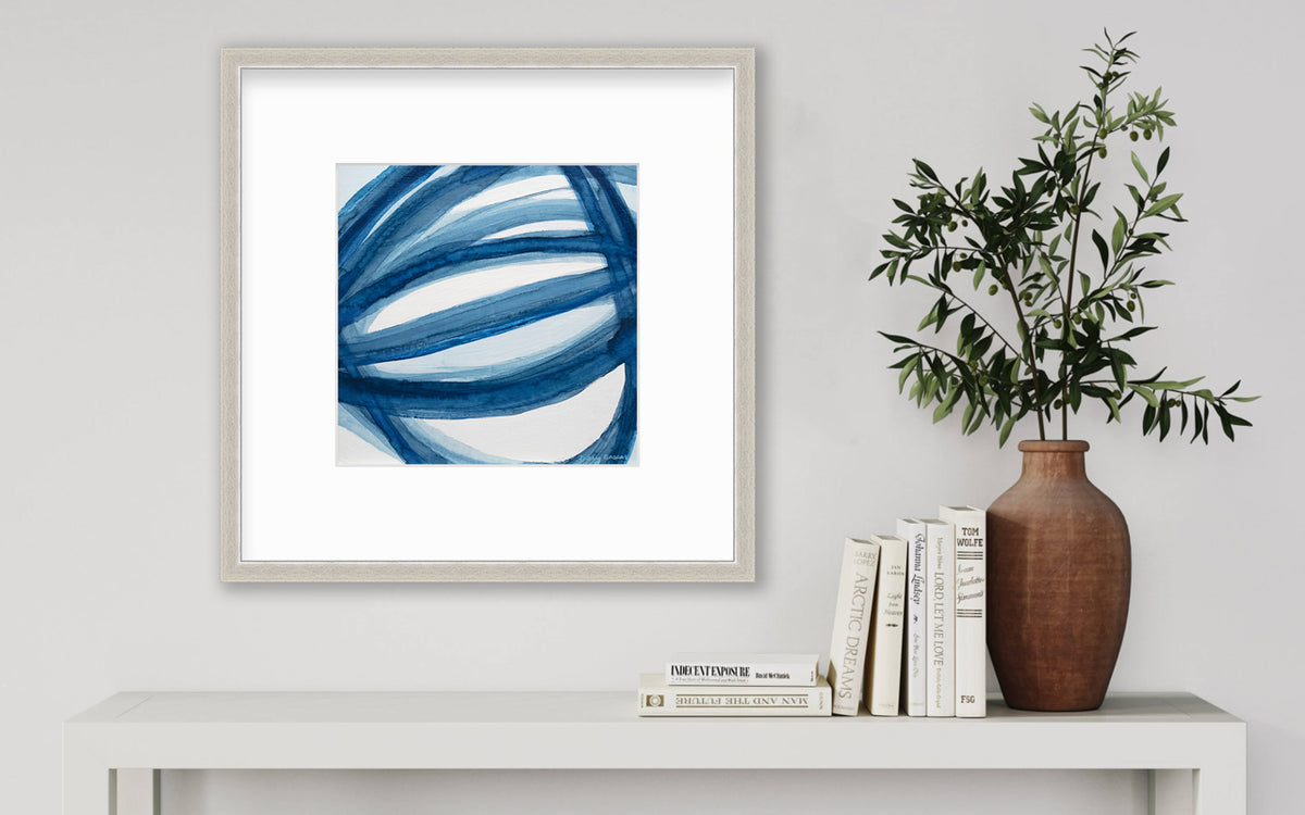 A blue and white abstract print in a silver frame with a mat hangs on a white wall above a white side table with a stack of books and a terracotta vase with greenery stems.