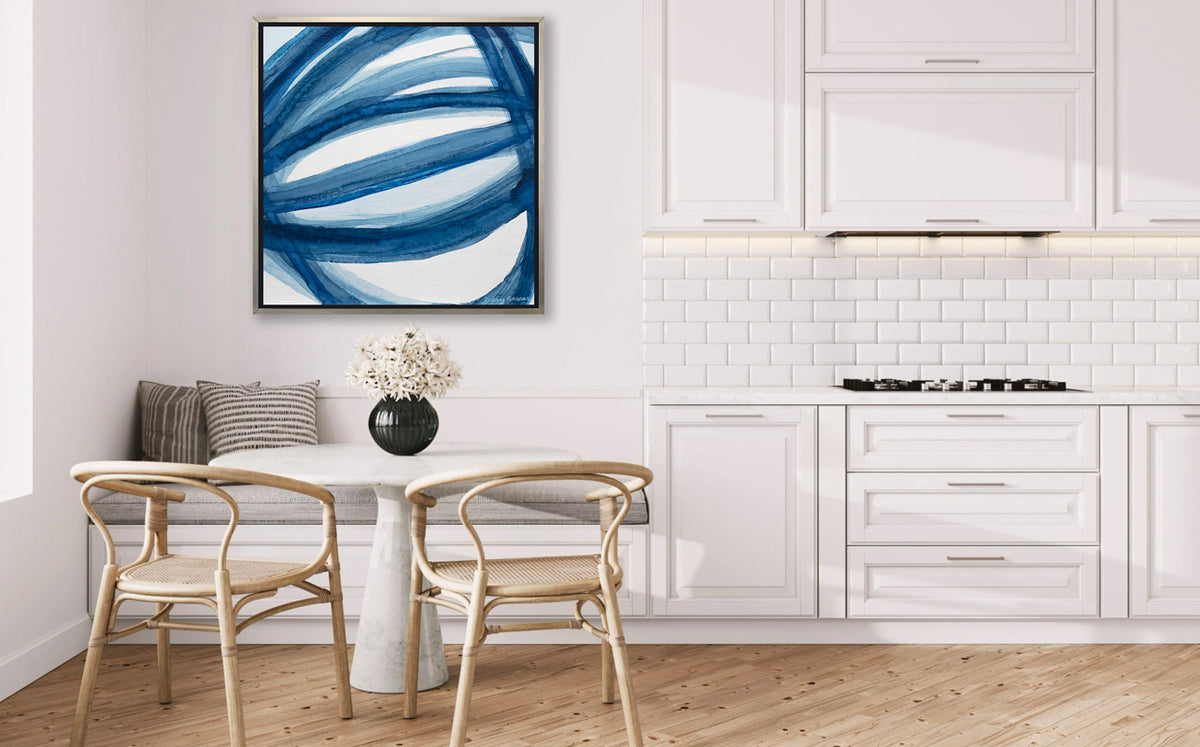 A blue and white abstract print in a silver floater frame hangs on a white wall above a white booth with a grey cushion, a white marble pedestal table, two natural wood chairs and next to white cabinets.