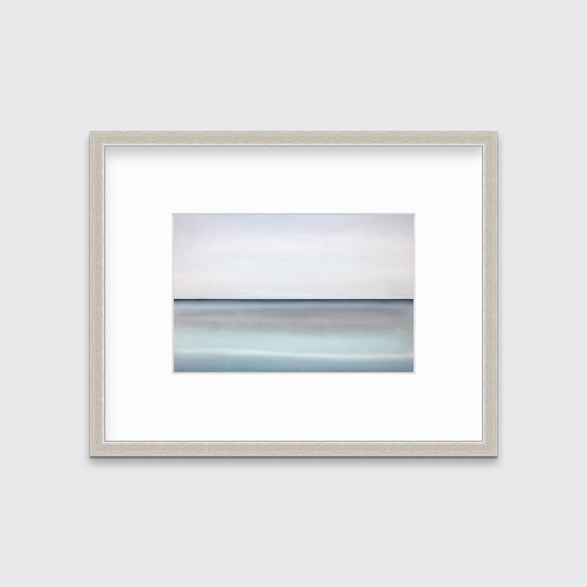 A tonal blue abstract landscape with a defined horizon print in a silver frame with a mat hangs on a white wall.