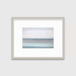 A tonal blue abstract landscape with a defined horizon print in a silver frame with a mat hangs on a white wall.