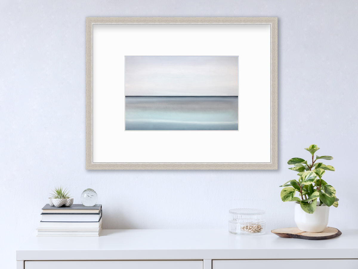 A tonal blue abstract landscape with a defined horizon print in a silver frame with a mat hangs on a white wall above a white surface with a stack of books, a clear round container with gold accessories and a potted plant.