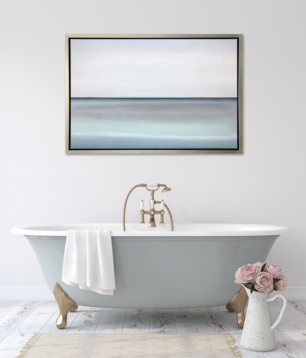 A tonal blue abstract landscape with a defined horizon print in a silver floater frame hangs on a white wall above a grey and white soaking bathtub with a white towel over an edge and a white pitcher filled with pink roses on the floor.
