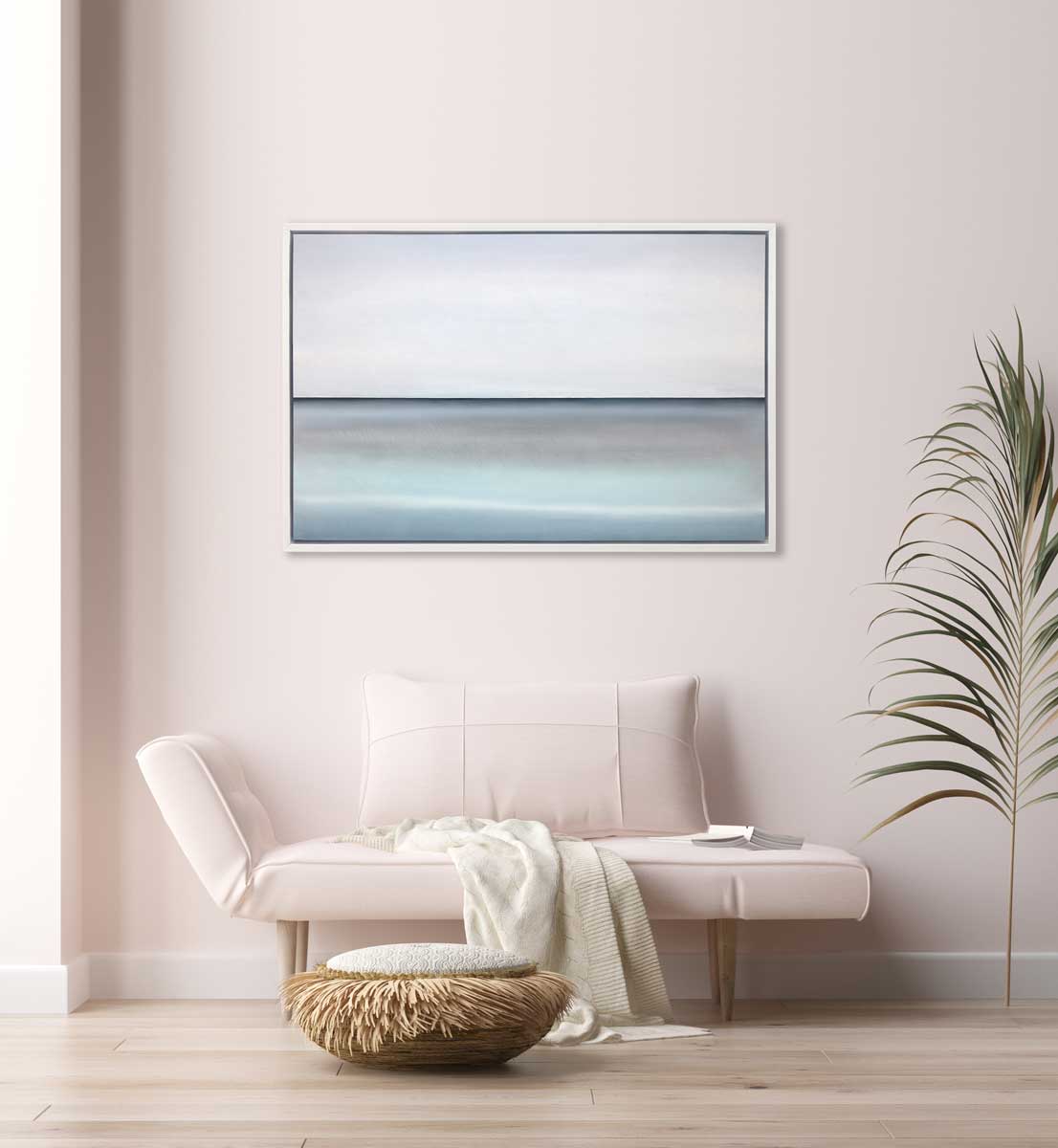 A tonal blue abstract landscape with a defined horizon print in a white floater frame hangs on a white wall above a light pink bench with a pillow, a white throw blanket and an open book. 