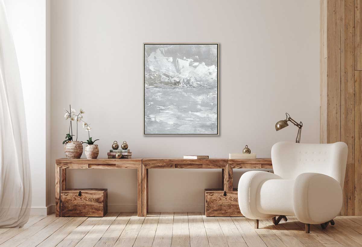 A white and grey abstract print in a silver floater frame hangs on a white wall above a wooden console table with two potted white orchids, a stack of books and a table lamp, a white modern chair is off to the right side.