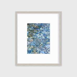 Abstracted river rocks under water hangs in a silver frame with a mat on a white wall.