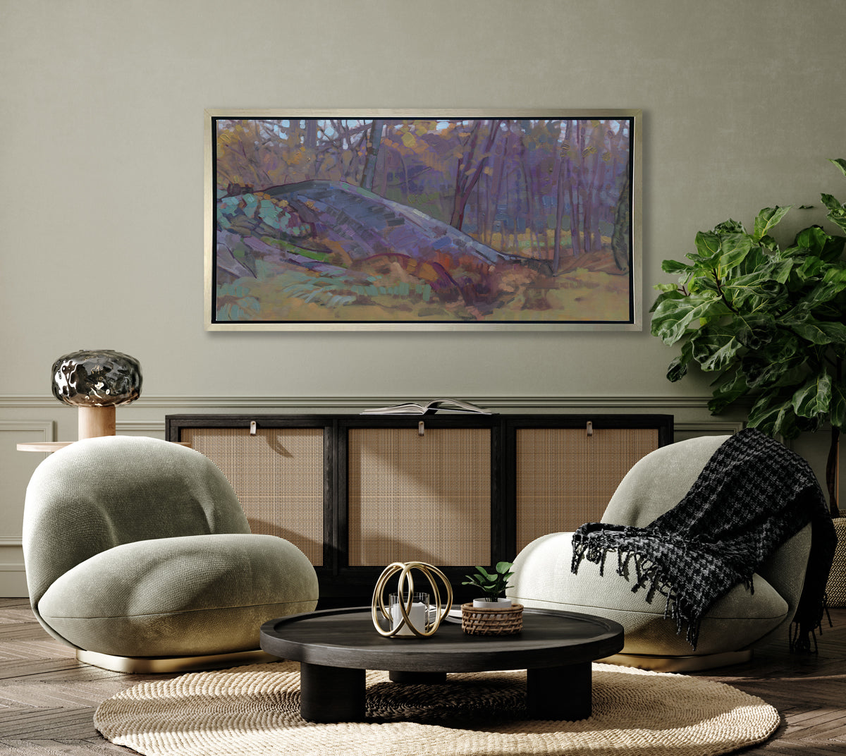 A muted multicolored abstract landscape print in a silver floater frame hangs on a beige wall above a dark wood and rattan sideboard with an open book, two matching green suede chairs with a black throw blanket and a low round black coffee table with decorative accessories.