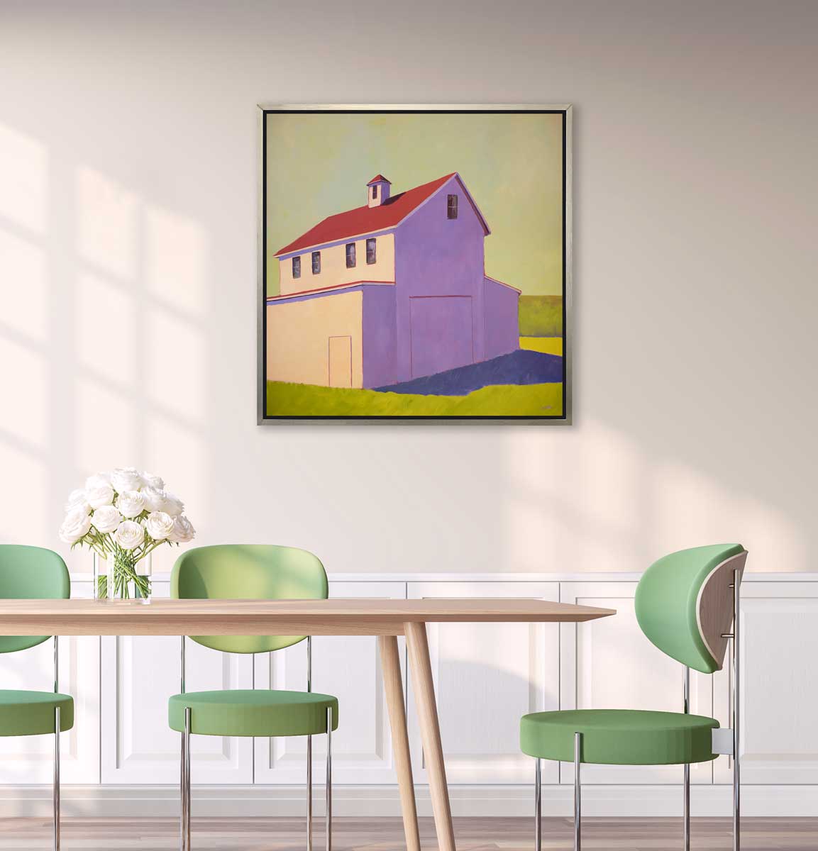 A lavender, green, beige and red contemporary barn print in a silver floater frame hangs on a white wall above a natural wood table with a glass vase of white roses and three light green chairs.