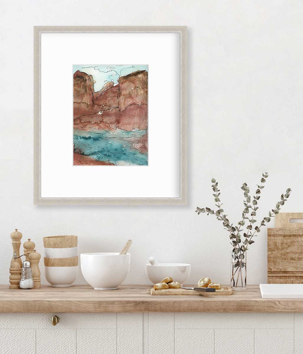 A teal, brown and black abstract landscape print in a silver frame with a mat hangs on a white wall above a butcher block countertop with bowls, a cutting board and a clear vase with eucalyptus stems.
