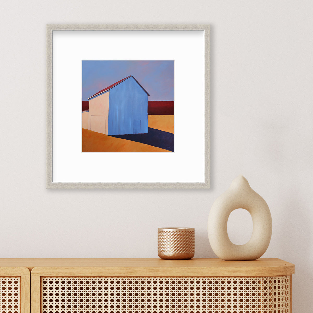 A lavender, red, beige and orange contemporary barn print in a silver frame with a mat hangs on a white wall above a natural wood cabinet with a copper candleholder and a white modern decorative accessory.
