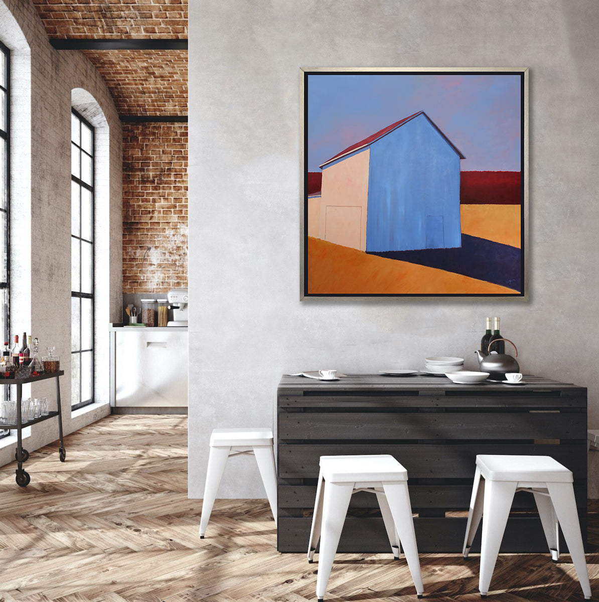 A lavender, red, beige and orange contemporary barn print in a silver floater frame hangs on a concrete wall above a black palette table with white place settings, four white stools and next to an opening to a well-lit hallway with a bar cart.