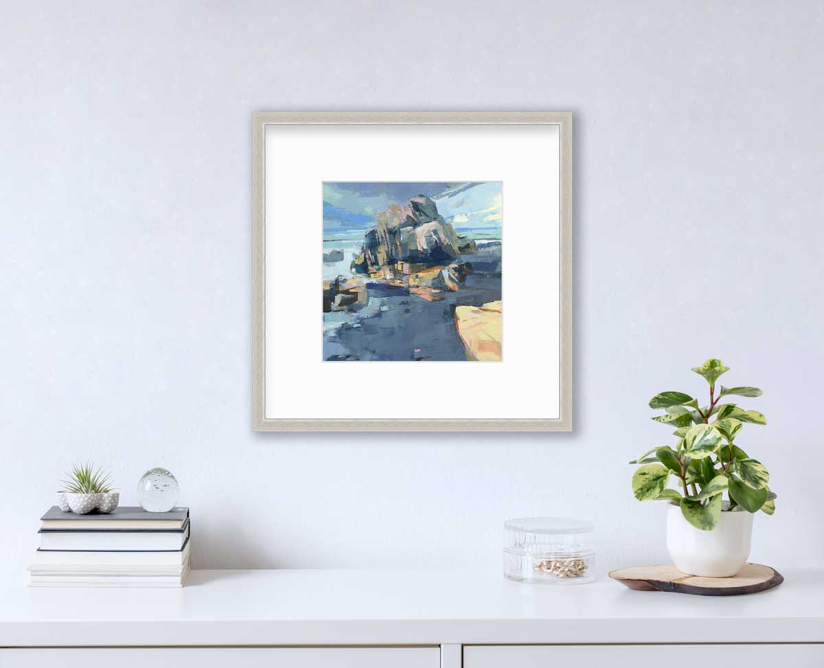 A blue, green and yellow abstract landscape print in a silver frame with a mat hangs on a white wall above a white surface with a stack of books, a round clear container with gold accessories and a potted plant.