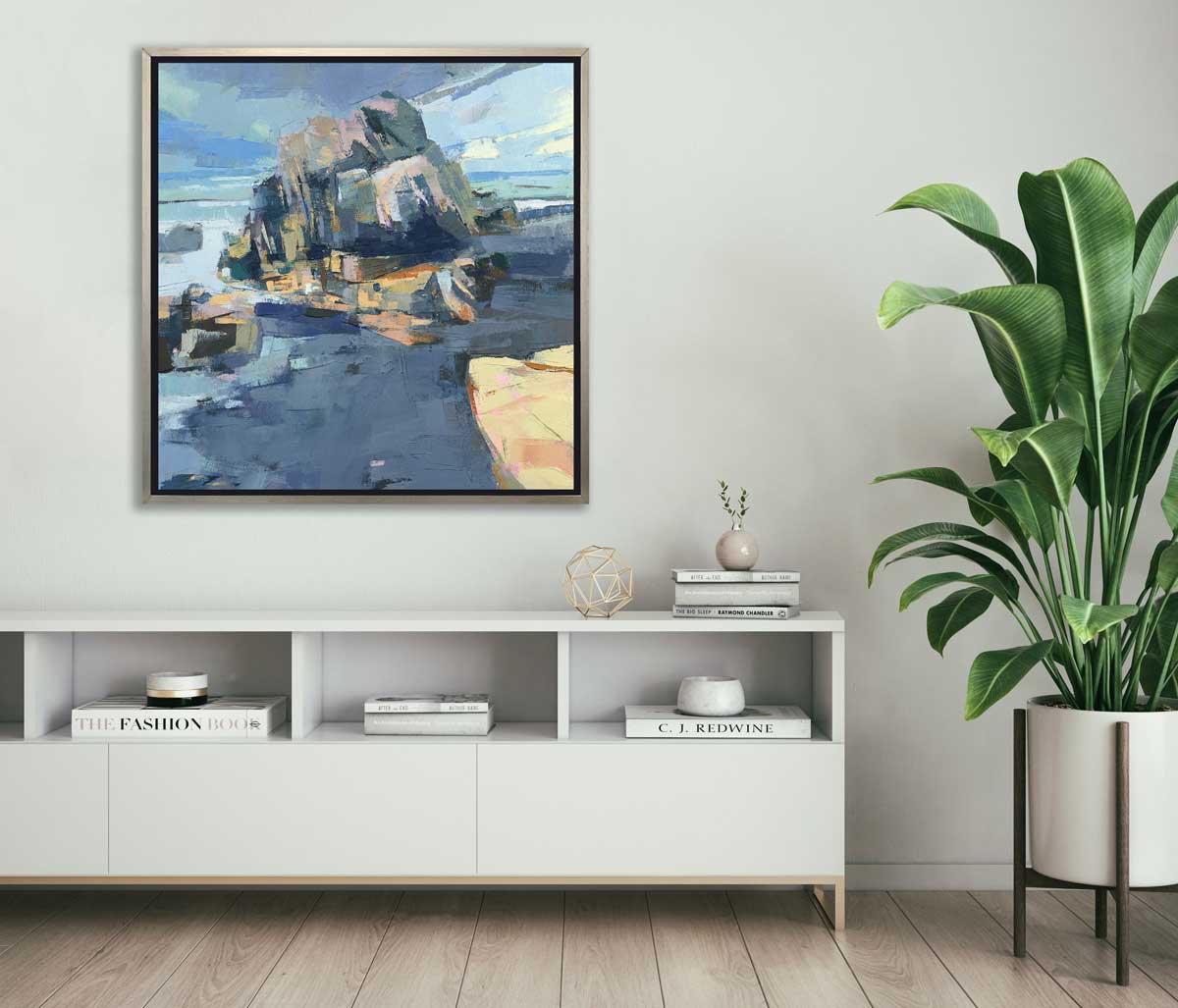 A blue, green and yellow abstract landscape print in a silver floater frame hangs on a white wall above a white shelf with stacks of books, a gold wire sphere and a potted floor plant.