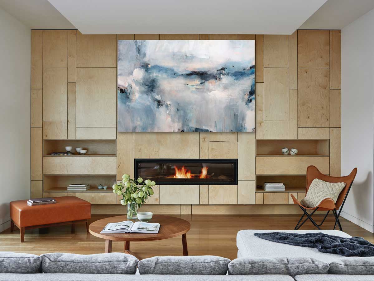 Cool toned abstract painting with spots of beige by Kelly Rossetti hangs on a geometric wood panel wall above a built-in fireplace and a grey sectional in front with a wood coffee table. 