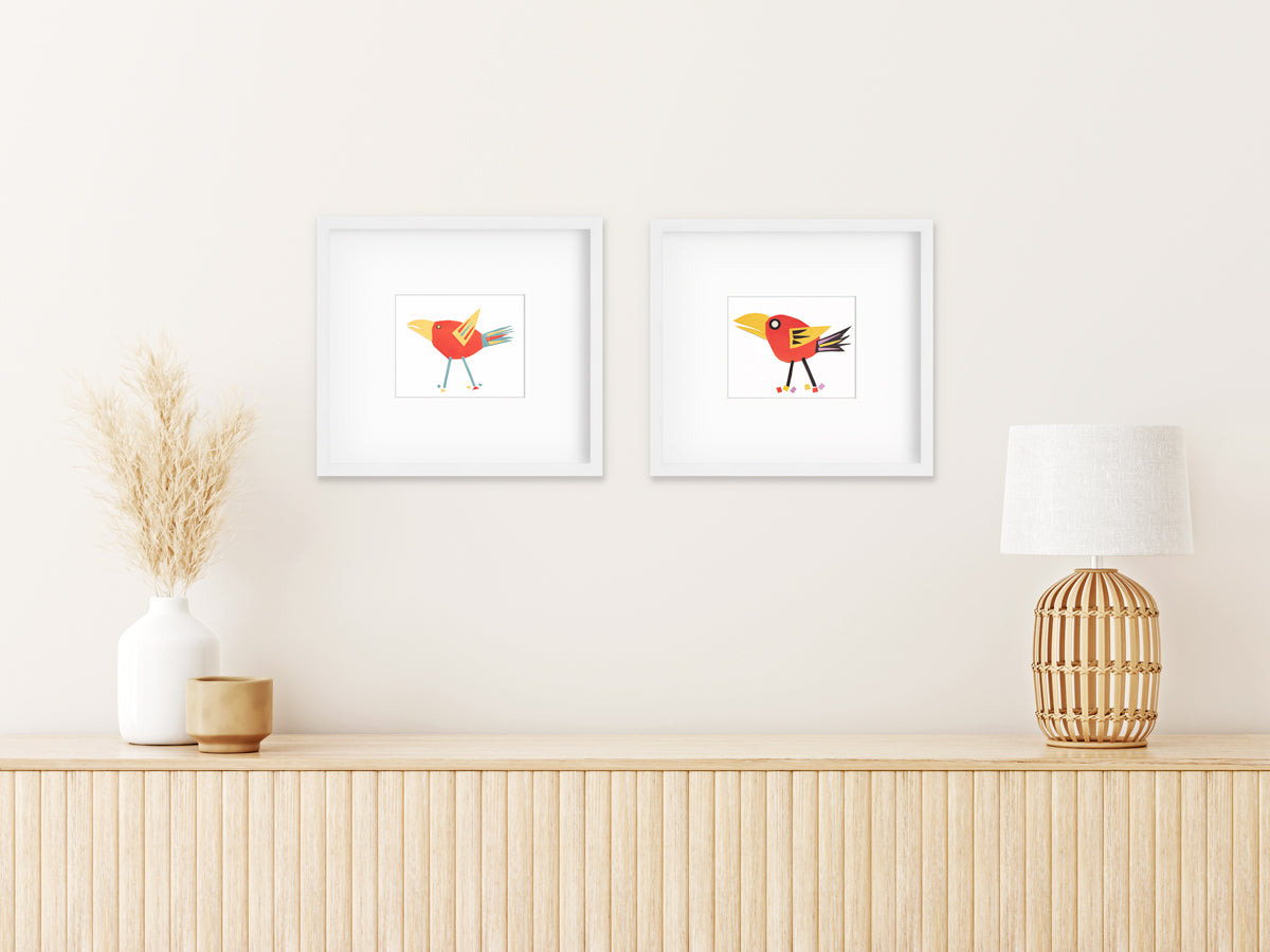 Two red bird collage prints in white frames with mats hang side by side on a white wall above a modern natural wood console with books and grey decorative vases on top.