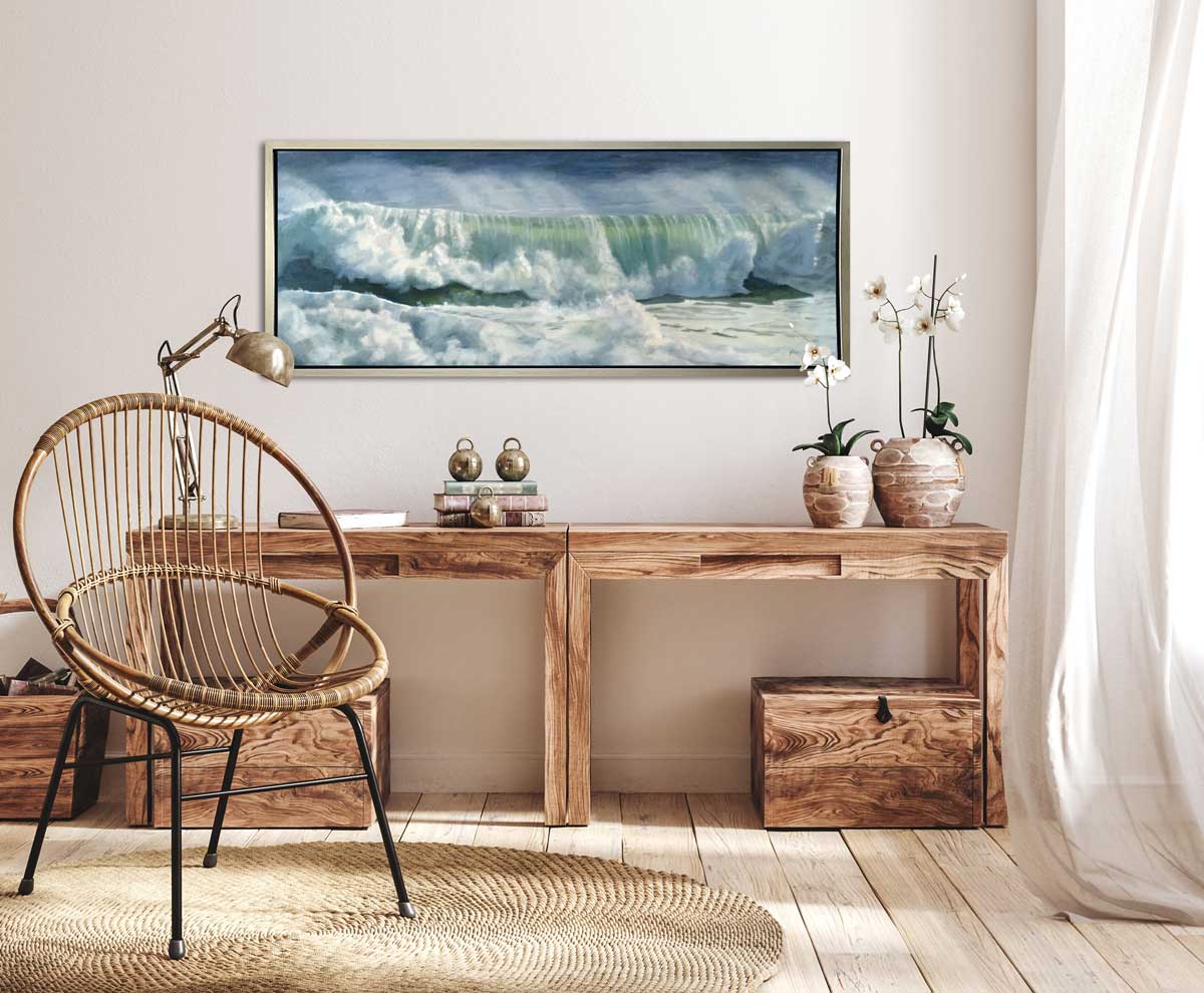 A blue, white and green abstract rolling wave coming to shore print in a silver floater frame hangs on a white wall above a wood elongated desk with two potted orchids, a stack of books, a desk lamp and a modern wire chair.