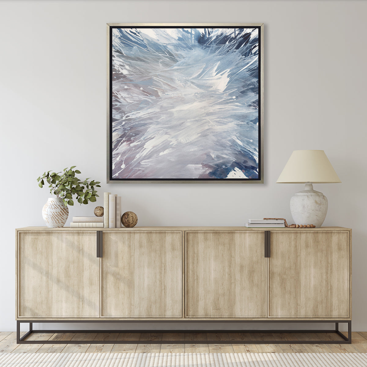 A light purple, grey, white and blue abstract print in a silver floater frame hangs on a white wall above a light wood credenza with a white table lamp, stacks of books and a white vase with eucalyptus leaves.