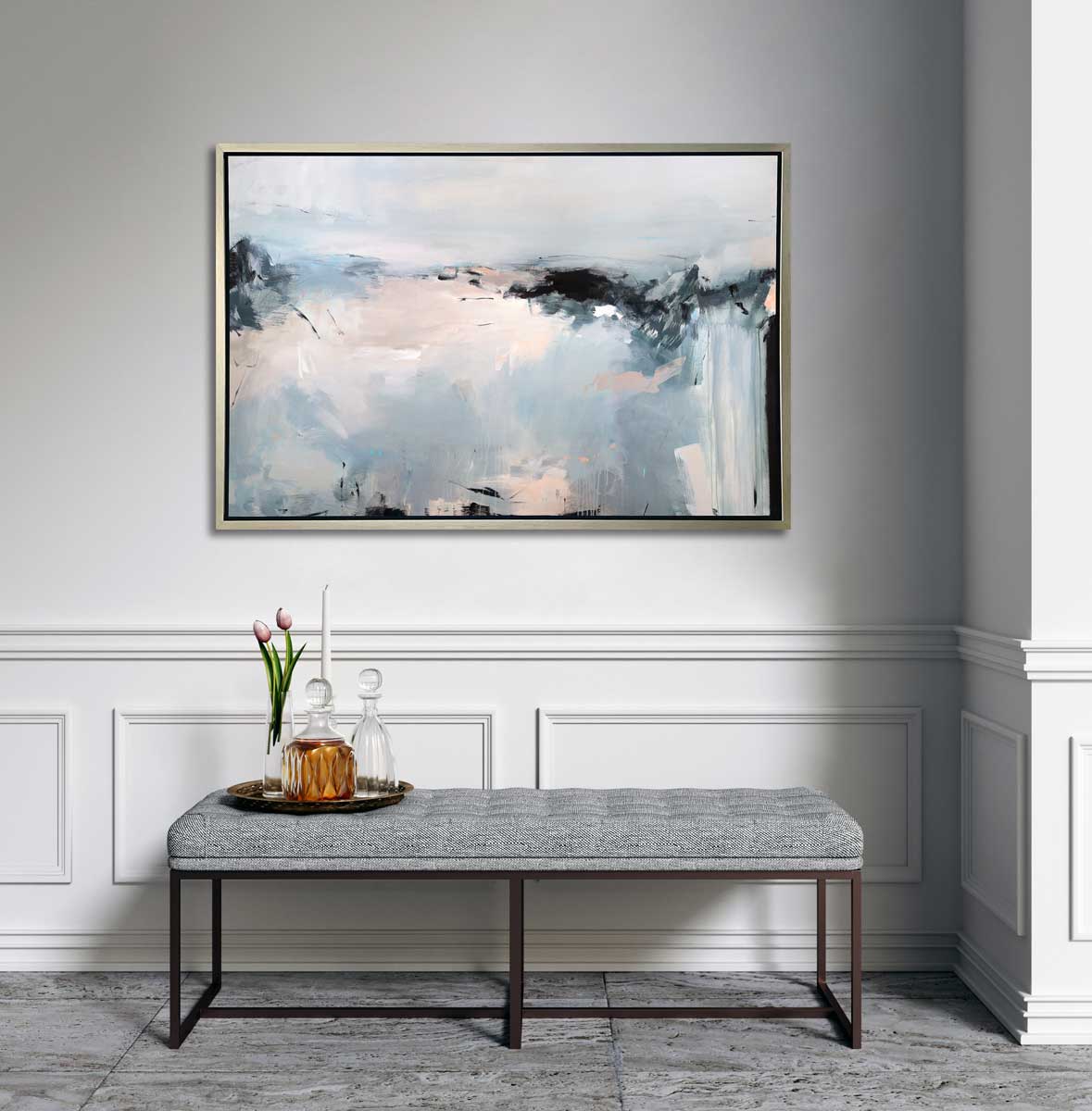 A light blue, white, light pink and black abstract print in a silver floater frame hangs on a white wall above a grey tufted bench with a tray of two glass decanters, a white tapered candle and a glass vase with two pink tulips.