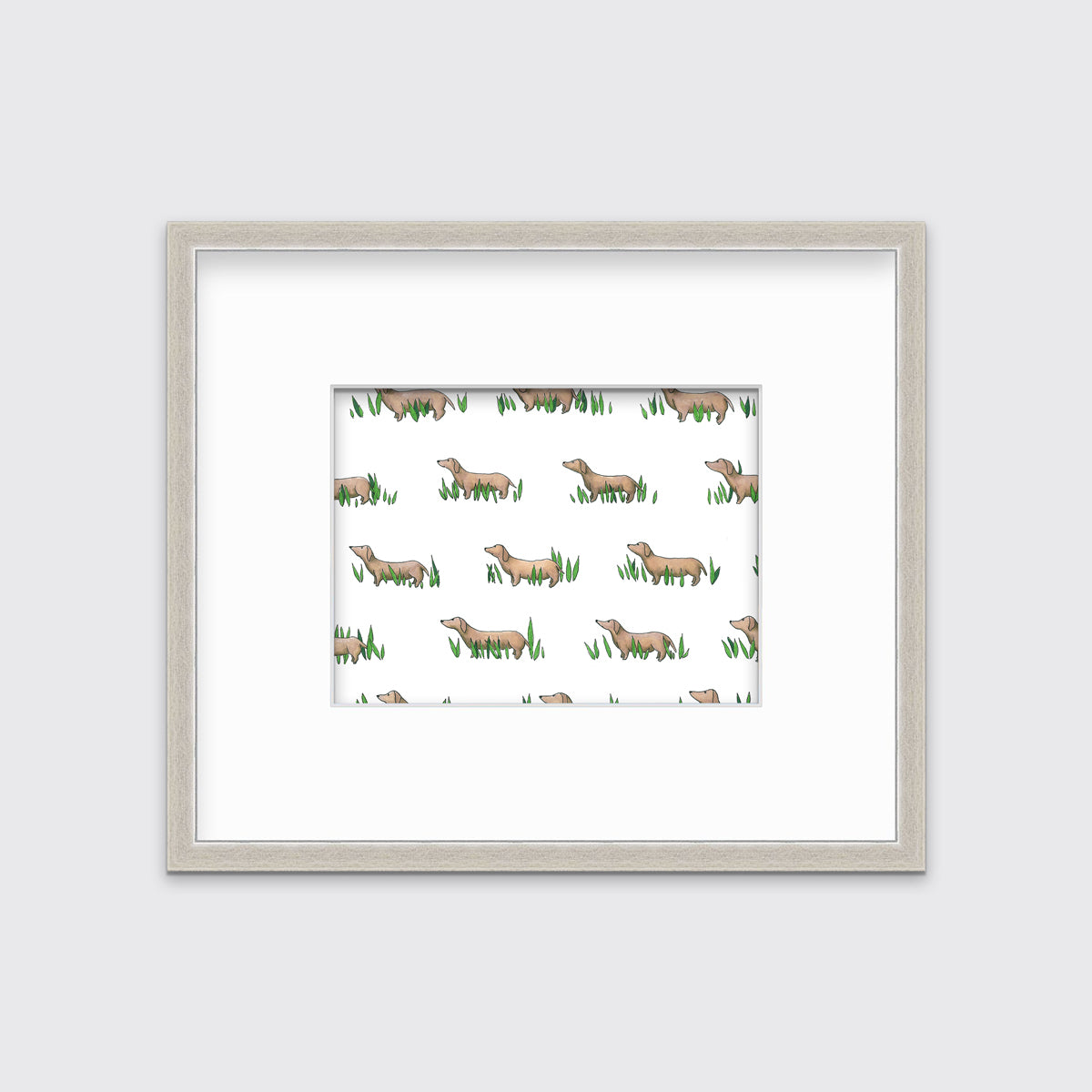 A print of brown dachshund dogs standing in green leaves in a silver frame with a mat hangs on a white wall.