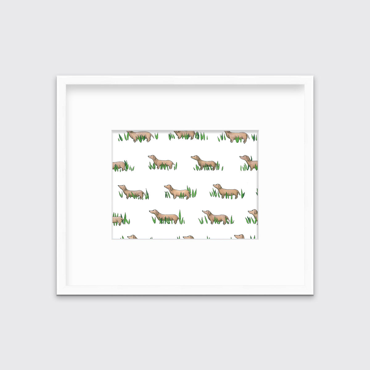 A print of brown dachshund dogs standing in green leaves in a white frame with a mat hangs on a white wall. 