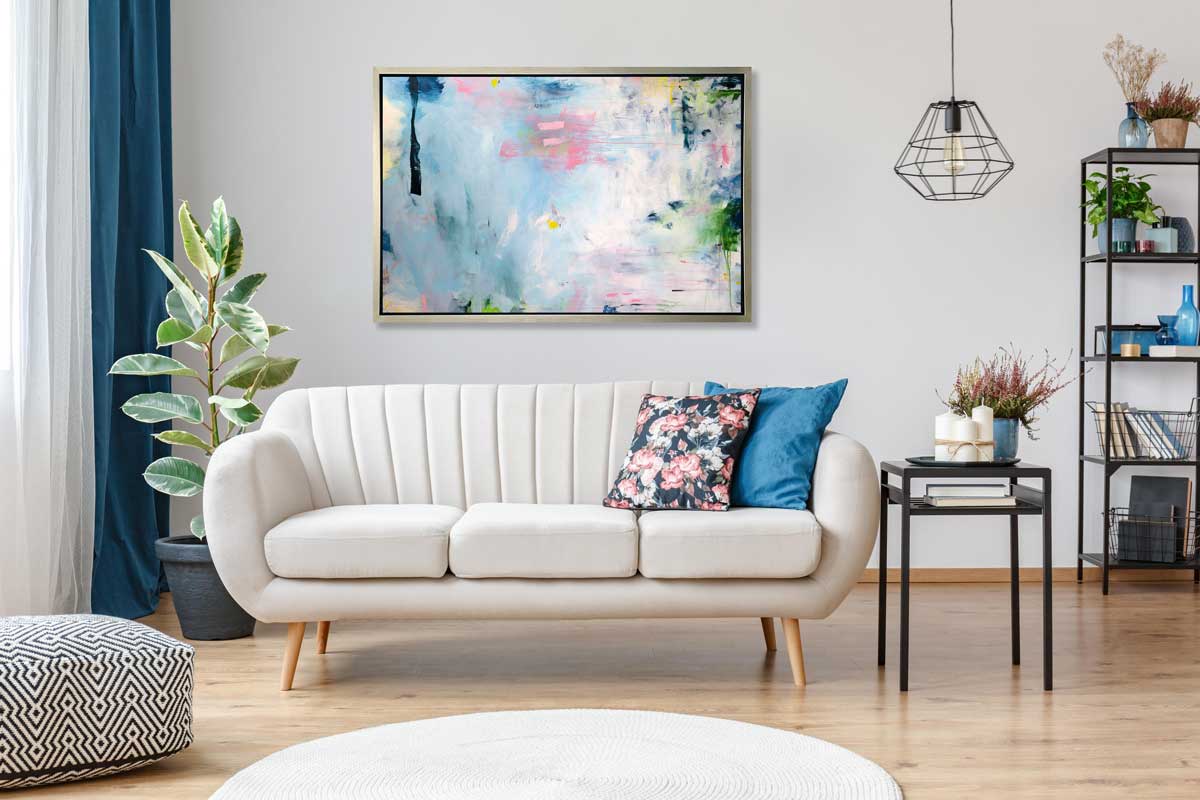 A blue, pink and white abstract print in a silver floater frame hangs on a white wall above a white sofa with pillows, a potted floor plant and a black side table with candles and a potted floral arrangement.