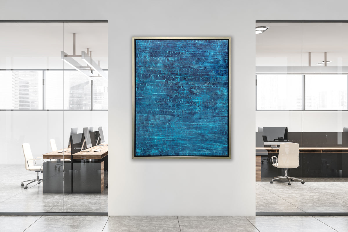 A dark teal abstract print in a silver floater frame hangs on a white wall in between two floor to ceiling windows to offices that have several desks, chairs and computer monitors.