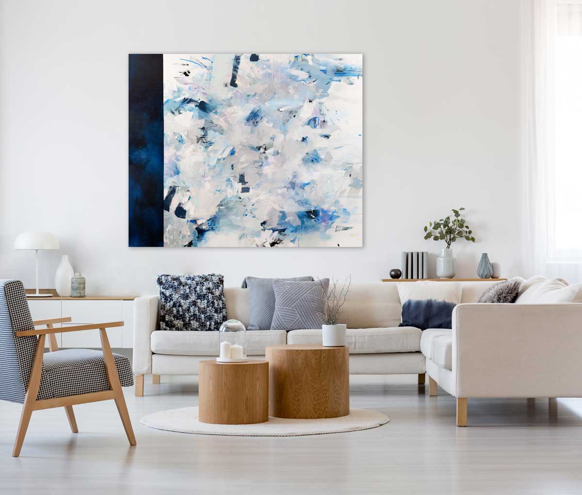A blue, white, and purple abstract painting with a navy blue thick line on the left side by Kelly Rossetti hangs on a white wall above beige sectional and two natural wood cylindrical coffee tables. The couch has blue and grey decor pillows and a mid century patterned arm chair is placed to the left of the sectional. 