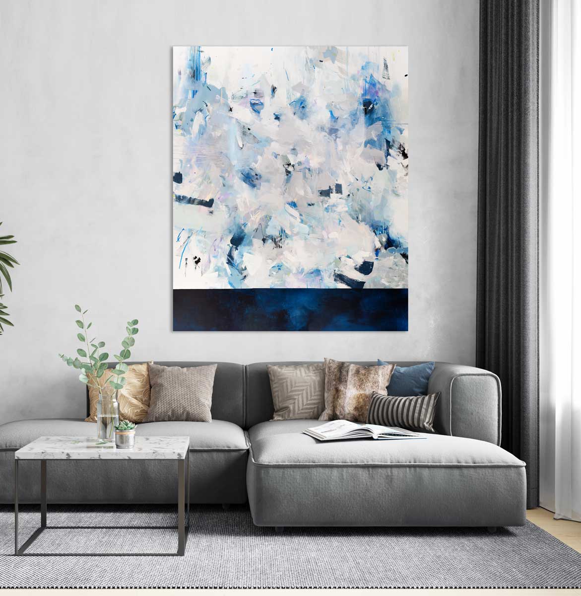 A blue, white, and purple abstract painting by Kelly Rossetti hangs vertically on a light grey wall above a modern grey sectional and a white marble coffee table with metal legs. 