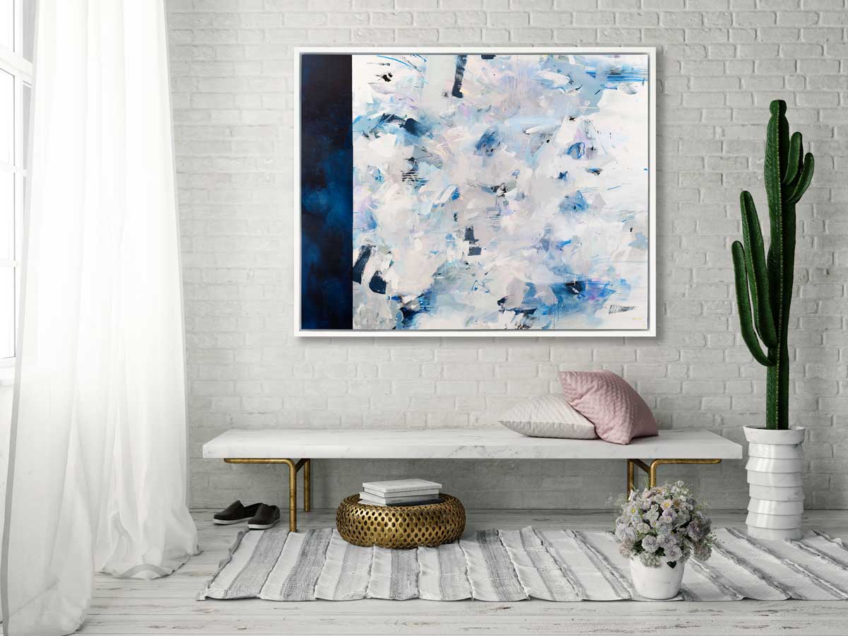 A blue, white and light grey abstract print in a white floater frame hangs horizontally on a white brick wall above a white marble bench with pillows, a large potted cactus and a gold ottoman with a stack of books.