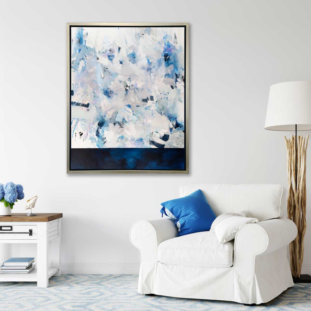 A blue, white and light grey abstract print in a silver floater frame hangs vertically on a white wall above a white overstuffed chair with a blue pillow, a driftwood floor lamp and a side table with a hydrangea arrangement, a shell decoration and a stack of books.