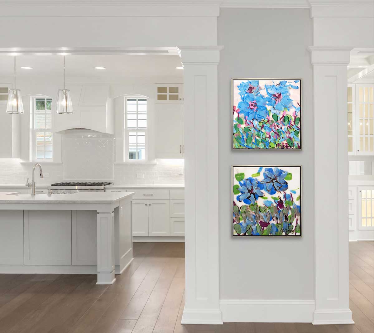 Two blue, green and white abstract floral prints in silver floater frames hang stacked on a light grey half wall next to a white kitchen.