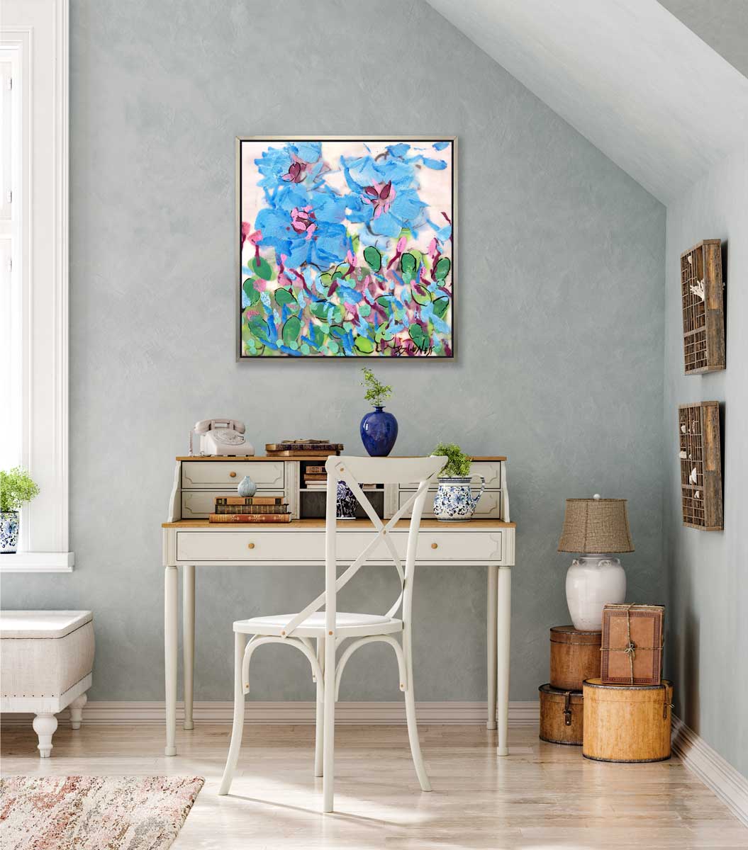 A blue, pink, green and white abstract floral print in a silver floater frame hangs on a light blue wall above a white two-tiered desk with a rotary phone, stacks of books, two vases with greenery and a white chair in front.