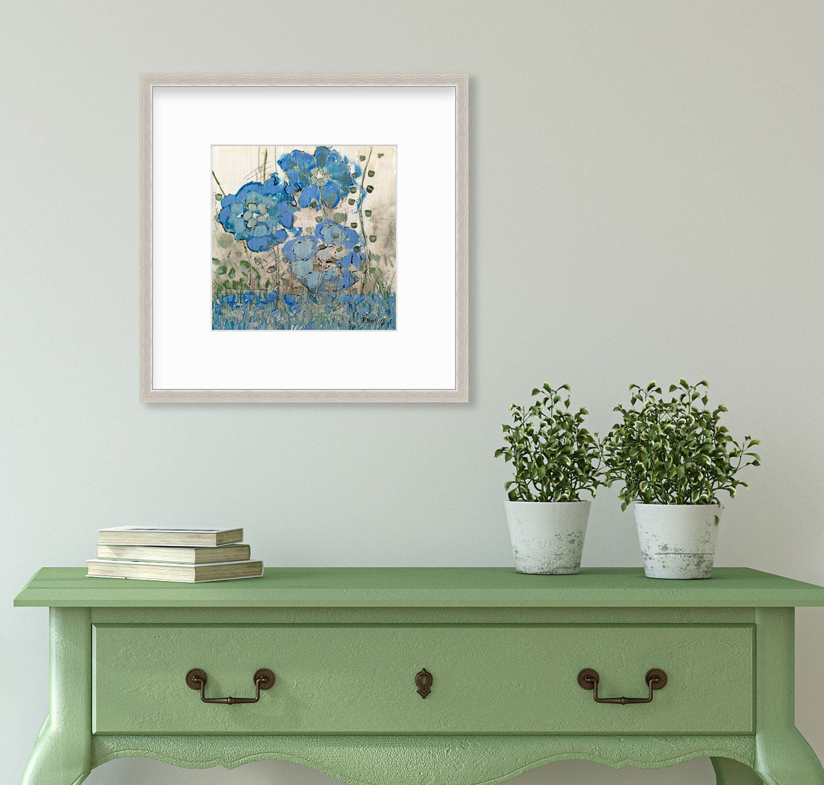 A blue floral print in a silver frame with a mat hangs on a white wall above a green console table with two potted plants and a stack of books.