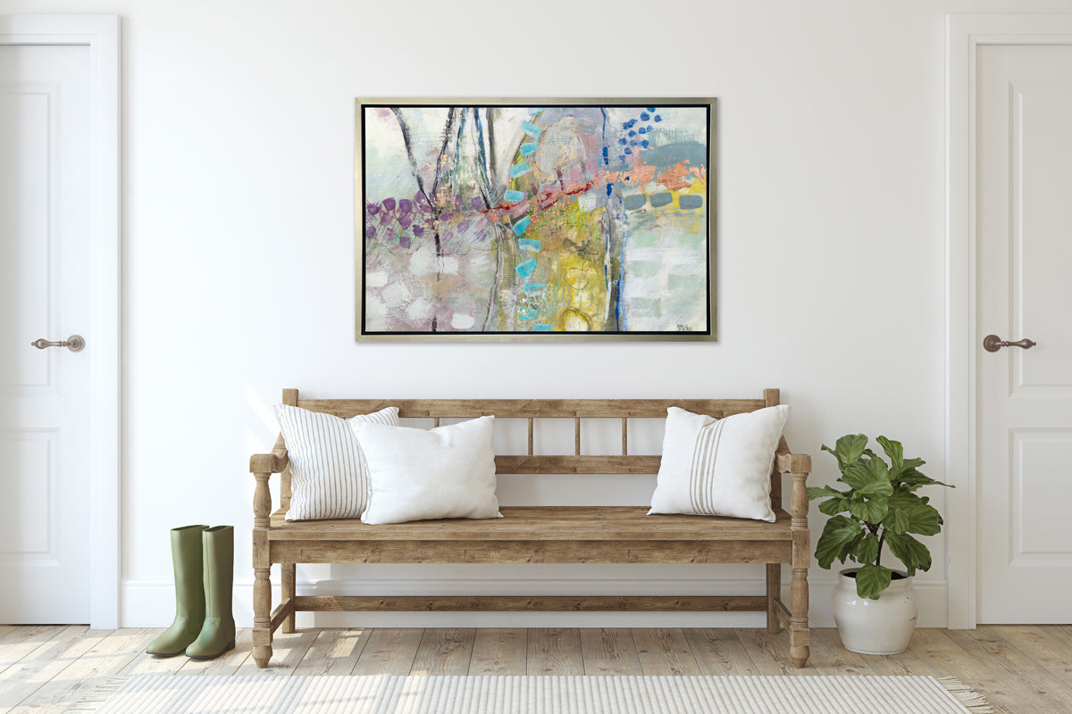 A multicolored abstract print in a silver floater frame hangs on a white wall above a whitewashed bench seat with pillows, a potted plant and a pair of green rubber boots.